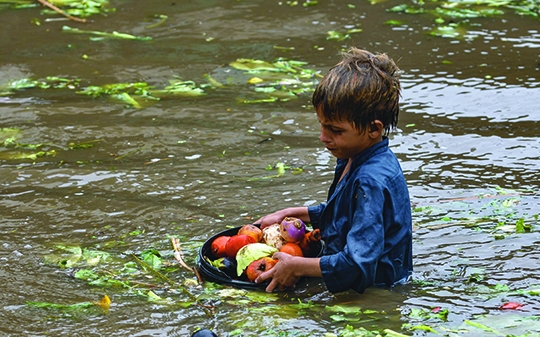 a small pakistani child wades waist deep in a flood with fruits in a basket
