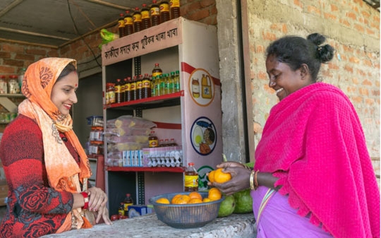 An Indian woman dressed with a pink shawl buys an orange from another lady in the shop